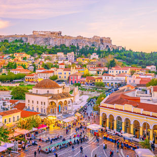 Druk Monastiraki-plein in Athene met terrassen en winkels, met op de achtergrond de Akropolis op een heuvel bij avondlicht.
